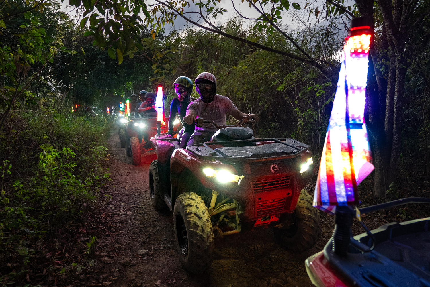 Two people on ATVs at night with lights on a trail surrounded by trees.